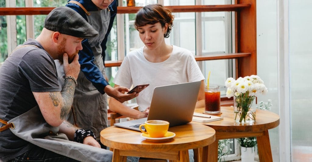 Three people at café table discussing phone, with laptop, coffee, and flowers
