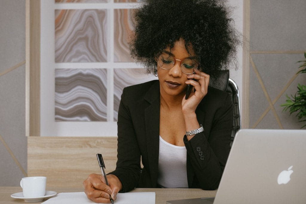 Woman with glasses on phone, writing notes at desk with laptop and plant