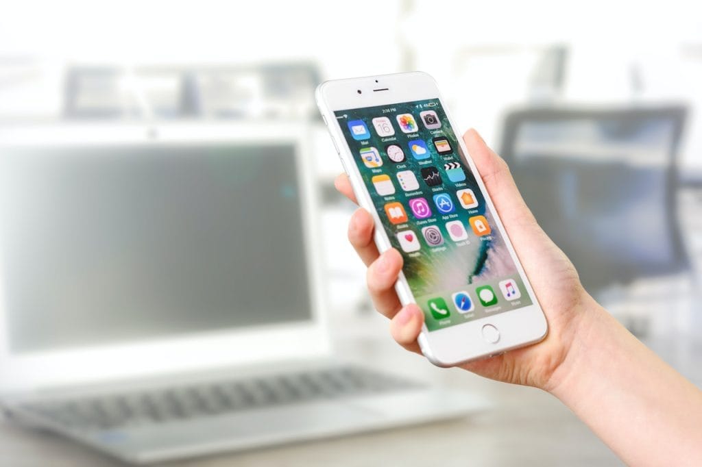 Hand holding smartphone with apps, blurred laptop in background workspace