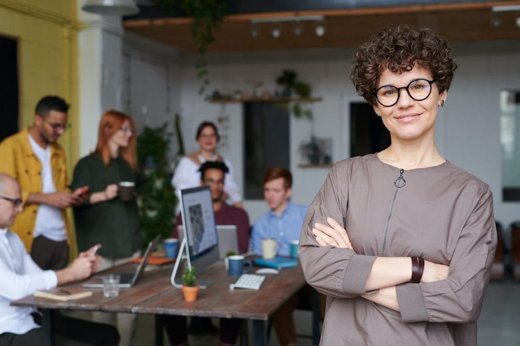 Confident woman with glasses standing before team working at table in office
