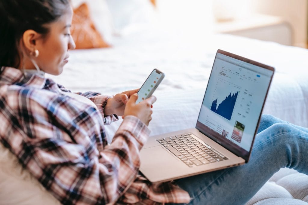 Woman using phone and laptop with financial charts