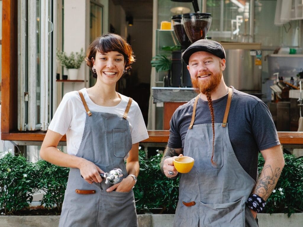 Two baristas smiling in café holding coffee tools